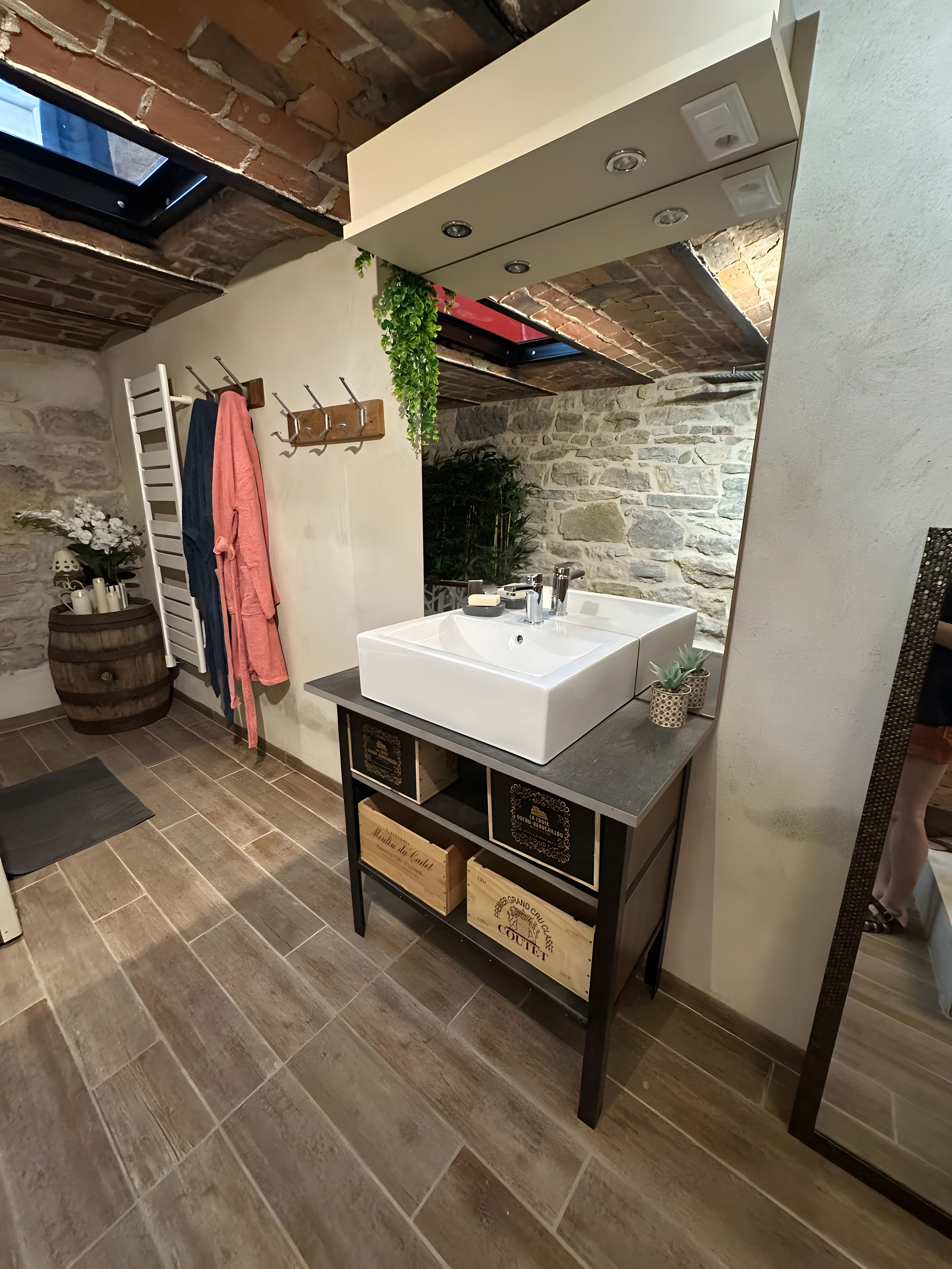 Une salle de bain avec un lavabo blanc, des meubles en bois, un porte-serviettes et un mur en pierre.
