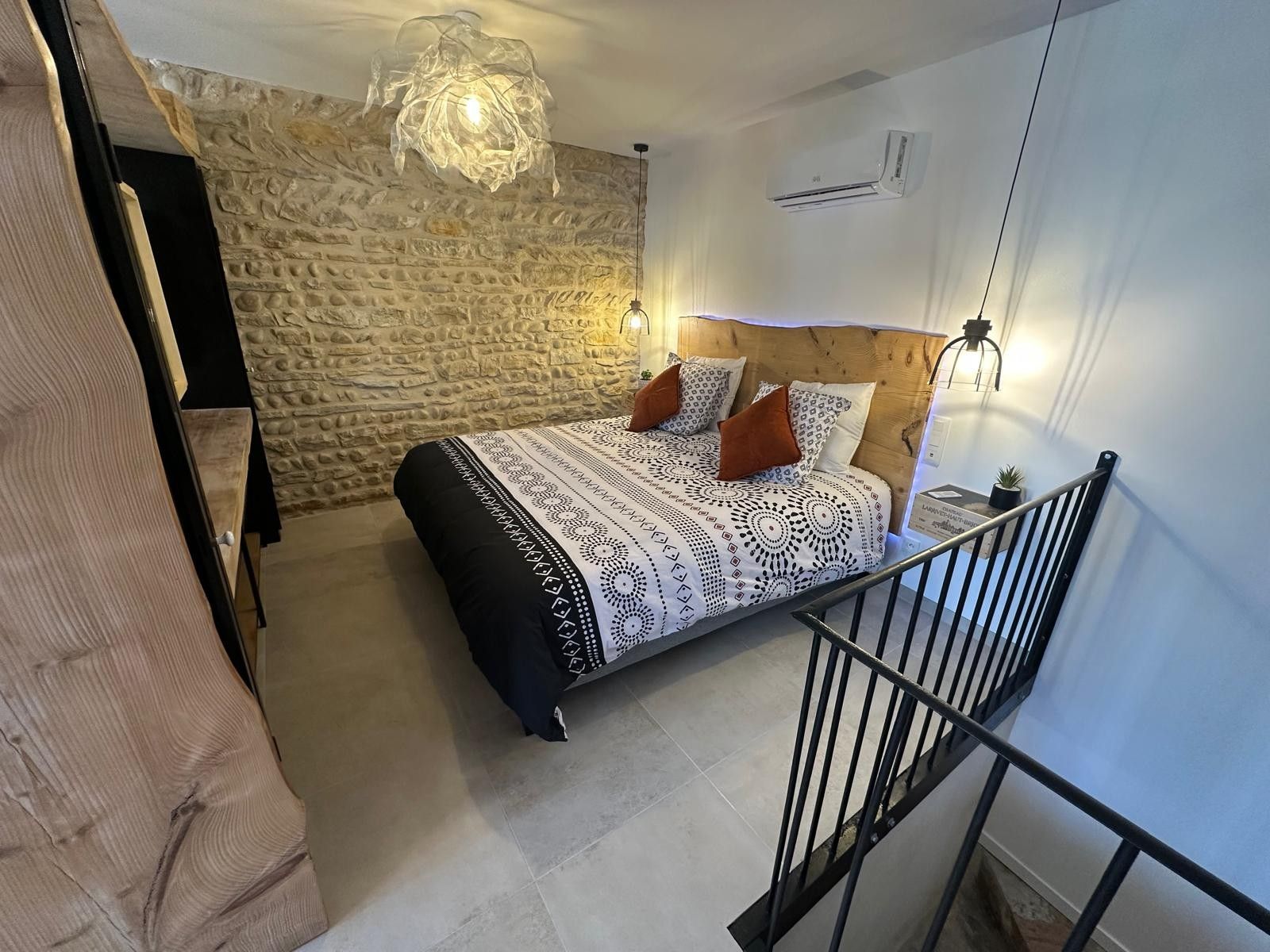Un dormitorio con pared de piedra, cabecera de madera, colcha estampada y lámpara colgante.