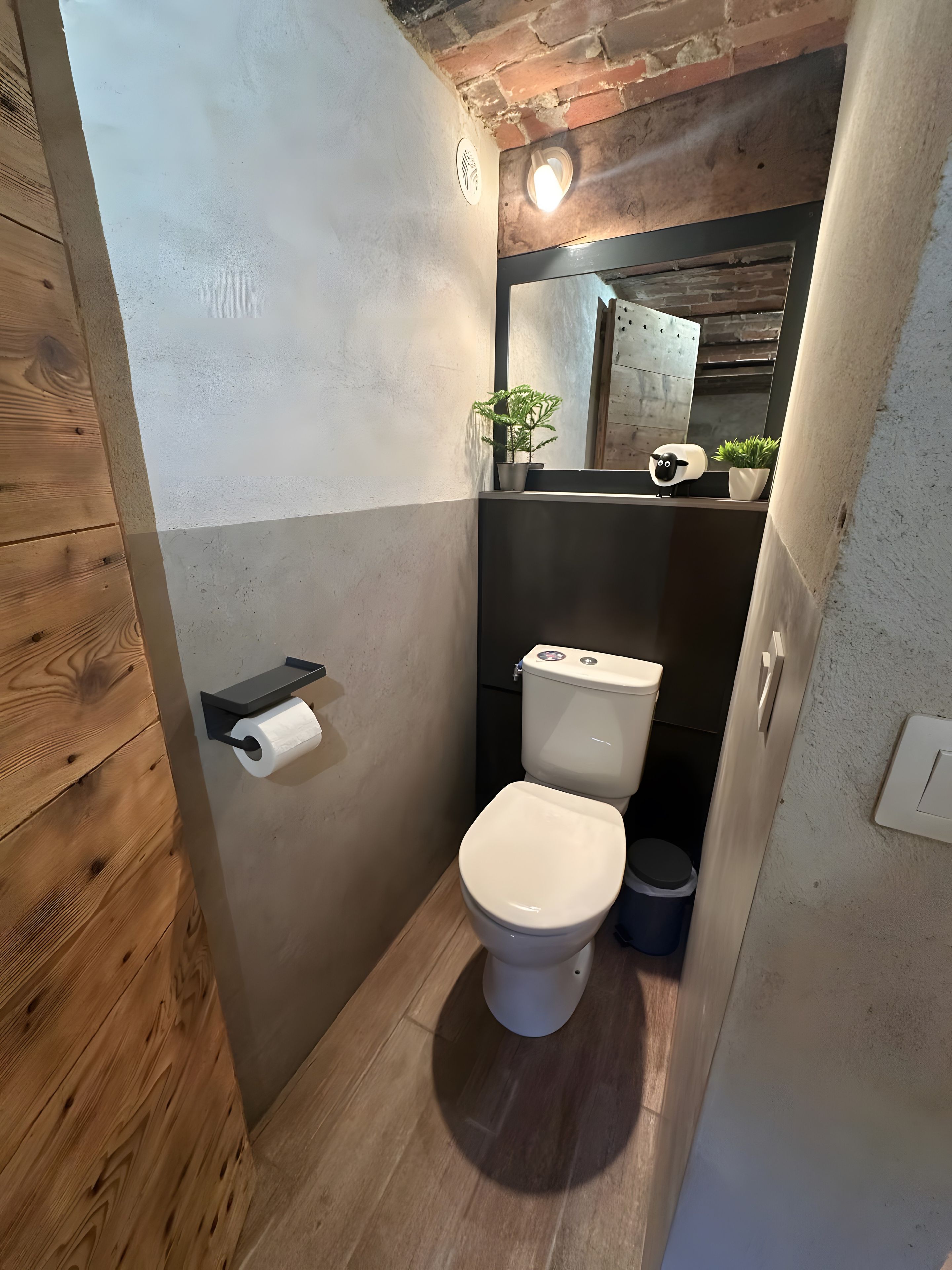 Een compacte badkamer met toilet, een klein prullenbakje en een plankje met een plant en een decoratie.