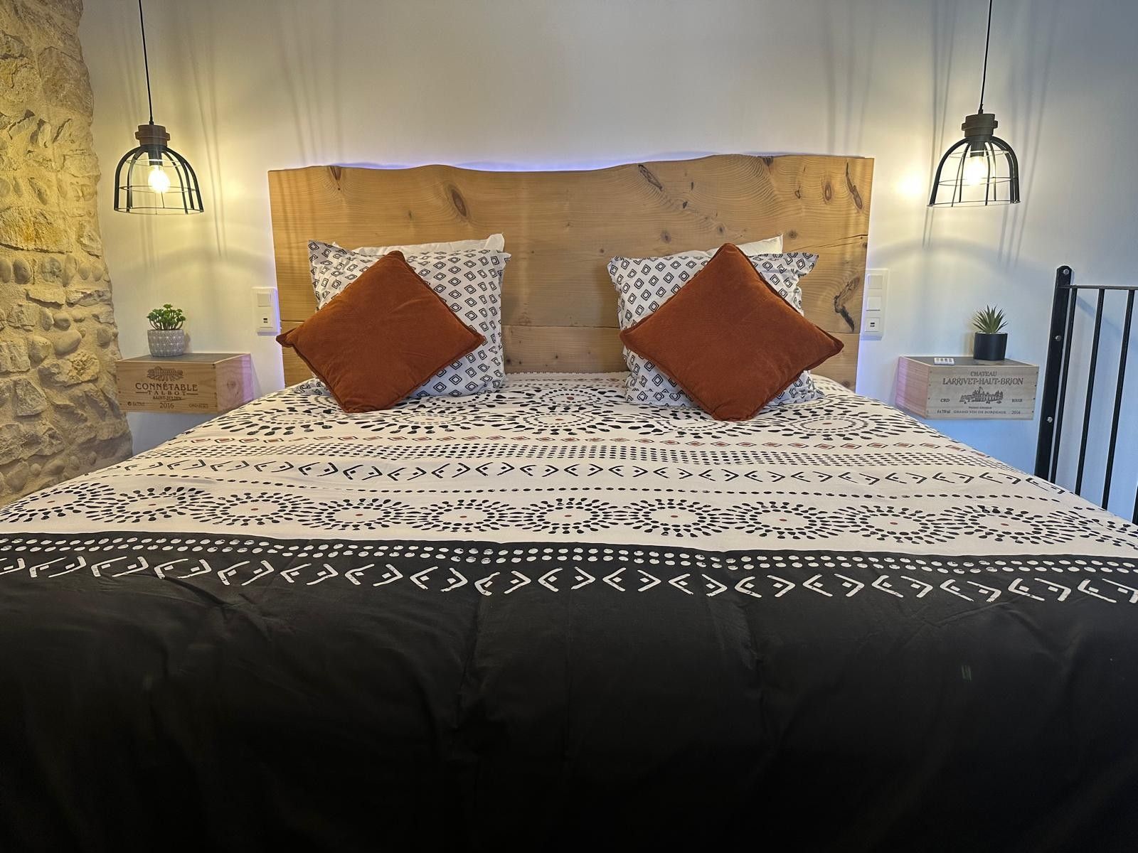 Un lit avec une literie à motifs, deux oreillers marron et une tête de lit en bois. Il y a deux tables de chevet avec de petites plantes et une table de nuit sur laquelle se trouve une boîte étiquetée « CONNÉTABLE ALBERT ».
