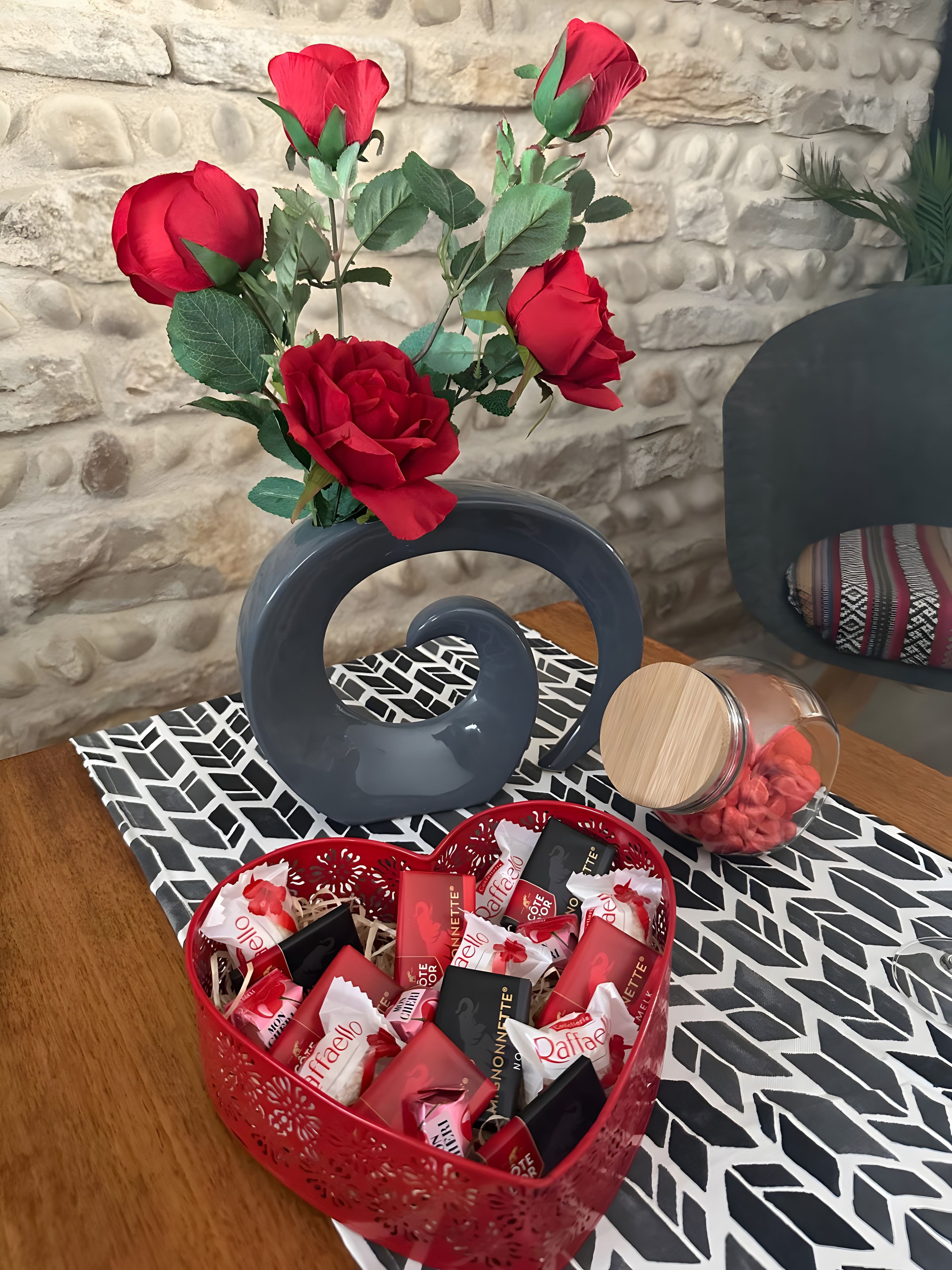 Un tavolo con un vaso di rose rosse, una ciotola decorativa e un cesto di cioccolatini.