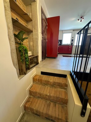 Una escalera con escalones de ladrillo que lleva a una puerta roja, una planta en maceta y vistas de una zona de cocina.