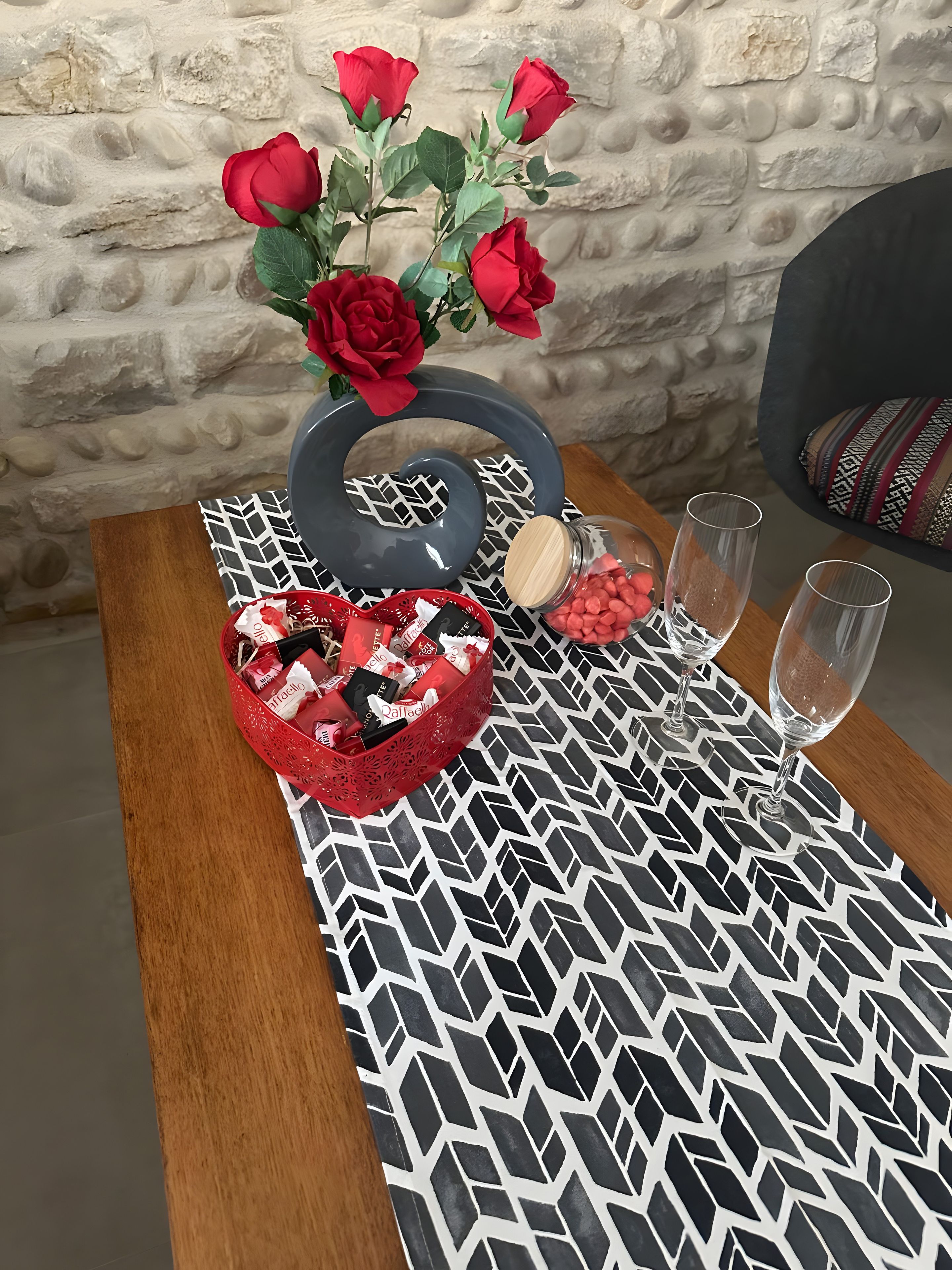 Una mesa con mantel estampado, un florero decorativo con rosas rojas, un objeto con forma de corazón, una cesta de bombones y dos vasos.