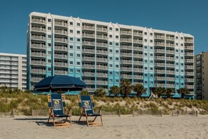 Hotel davant de la platja amb nombrosos balcons, sorra i dues cadies de platja amb una ombrel·la blava.