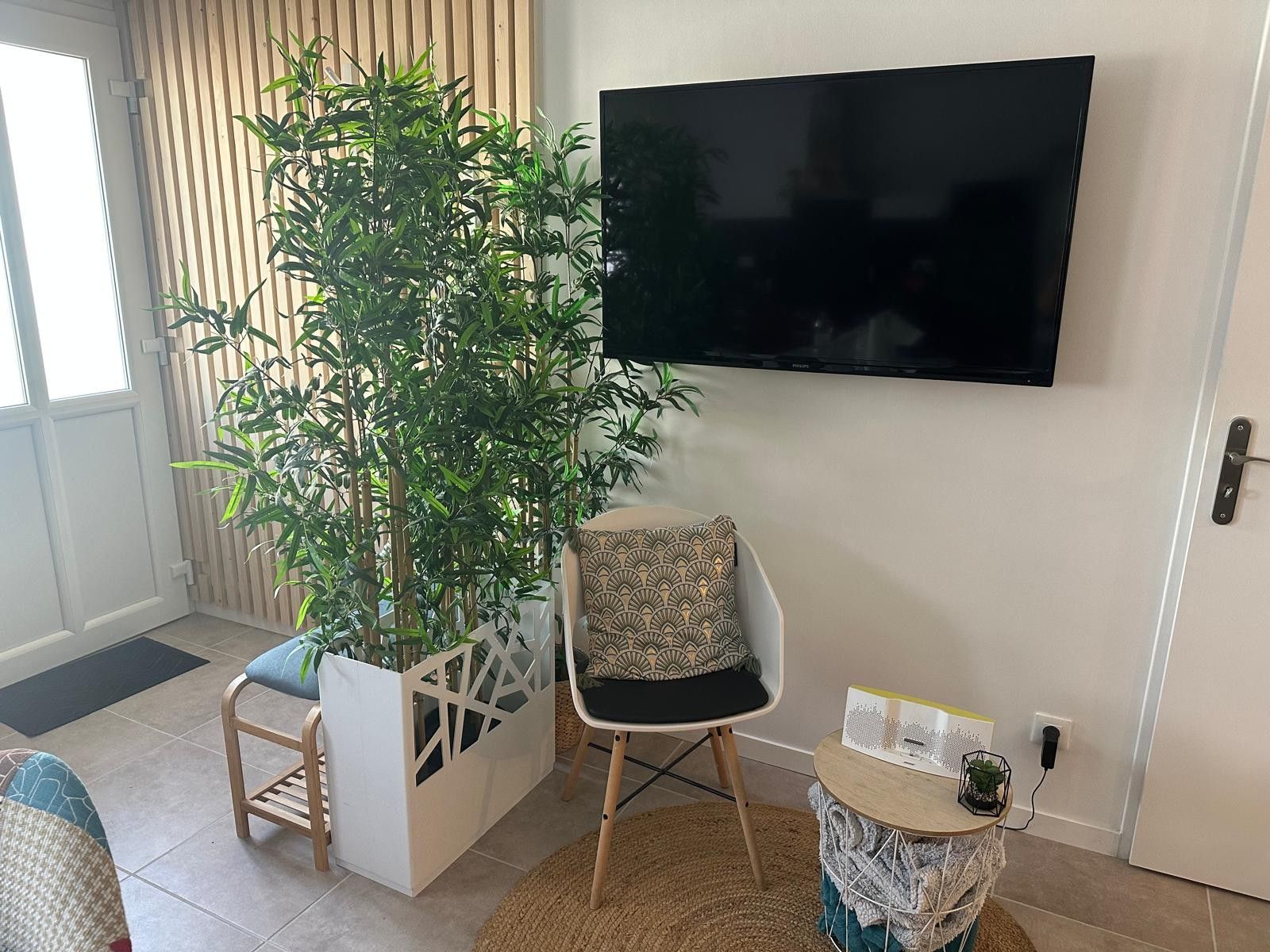 Un salon comprenant une grande plante en pot, un fauteuil moderne, une petite table ronde avec un livre et une plante, et un téléviseur à écran plat fixé au mur.
