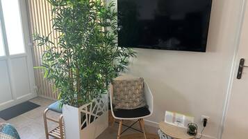Un salon comprenant une grande plante en pot, un fauteuil moderne, une petite table ronde avec un livre et une plante, et un téléviseur à écran plat fixé au mur.