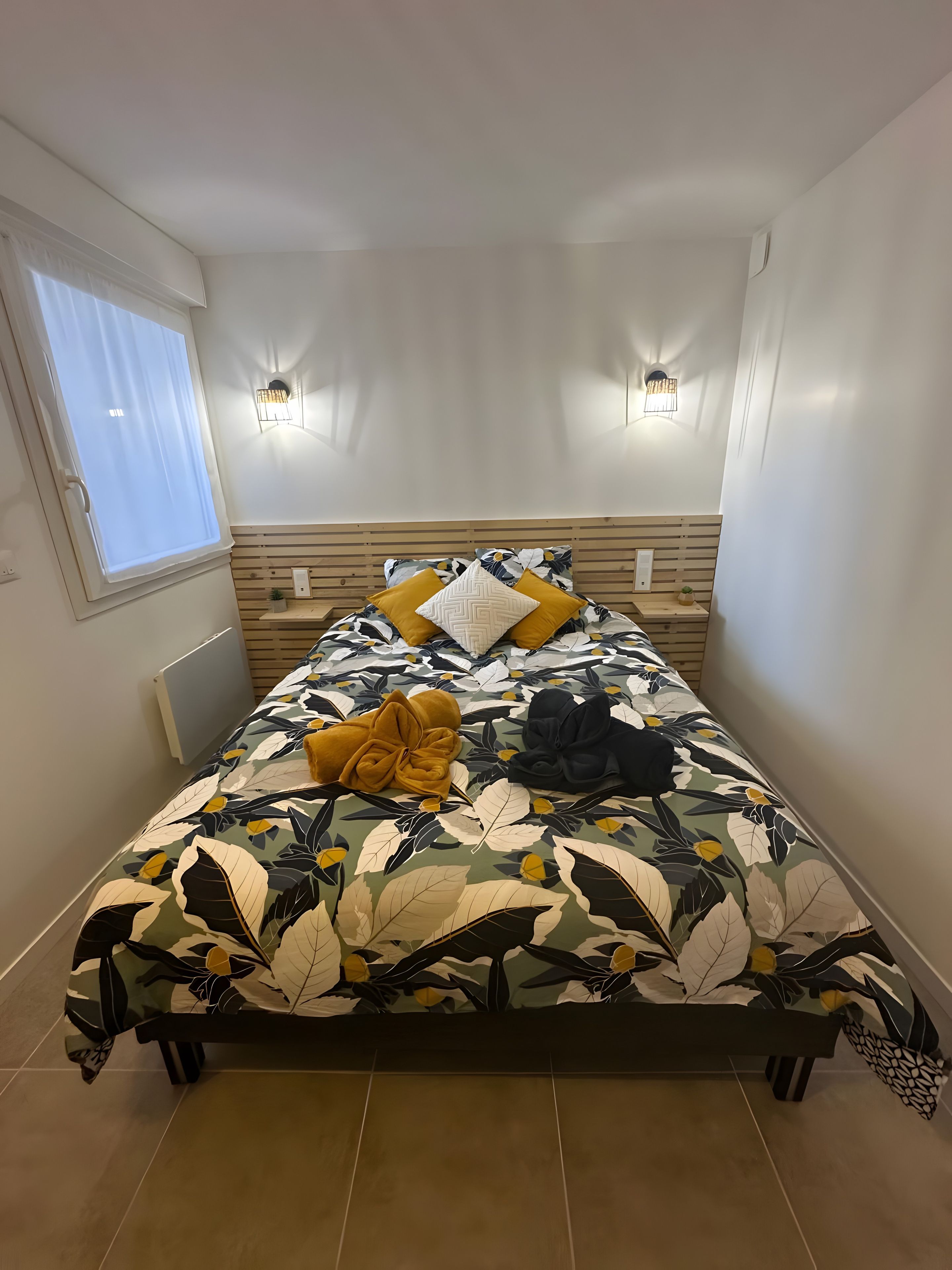 Un lit avec une couvre-lit à motifs floraux, deux lampes fixées au mur, une fenêtre avec des rideaux et un radiateur.