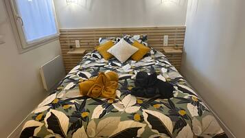 Un lit avec une couvre-lit à motifs floraux, deux lampes fixées au mur, une fenêtre avec des rideaux et un radiateur.