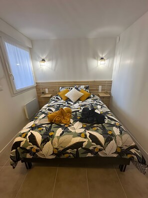 A bed with a floral bedspread, two wall-mounted lights, a window with curtains, and a radiator. - Gîte Le Méridi’Ain Apartment in Viriat: 1 Bedroom, Private Sauna (Viriat)