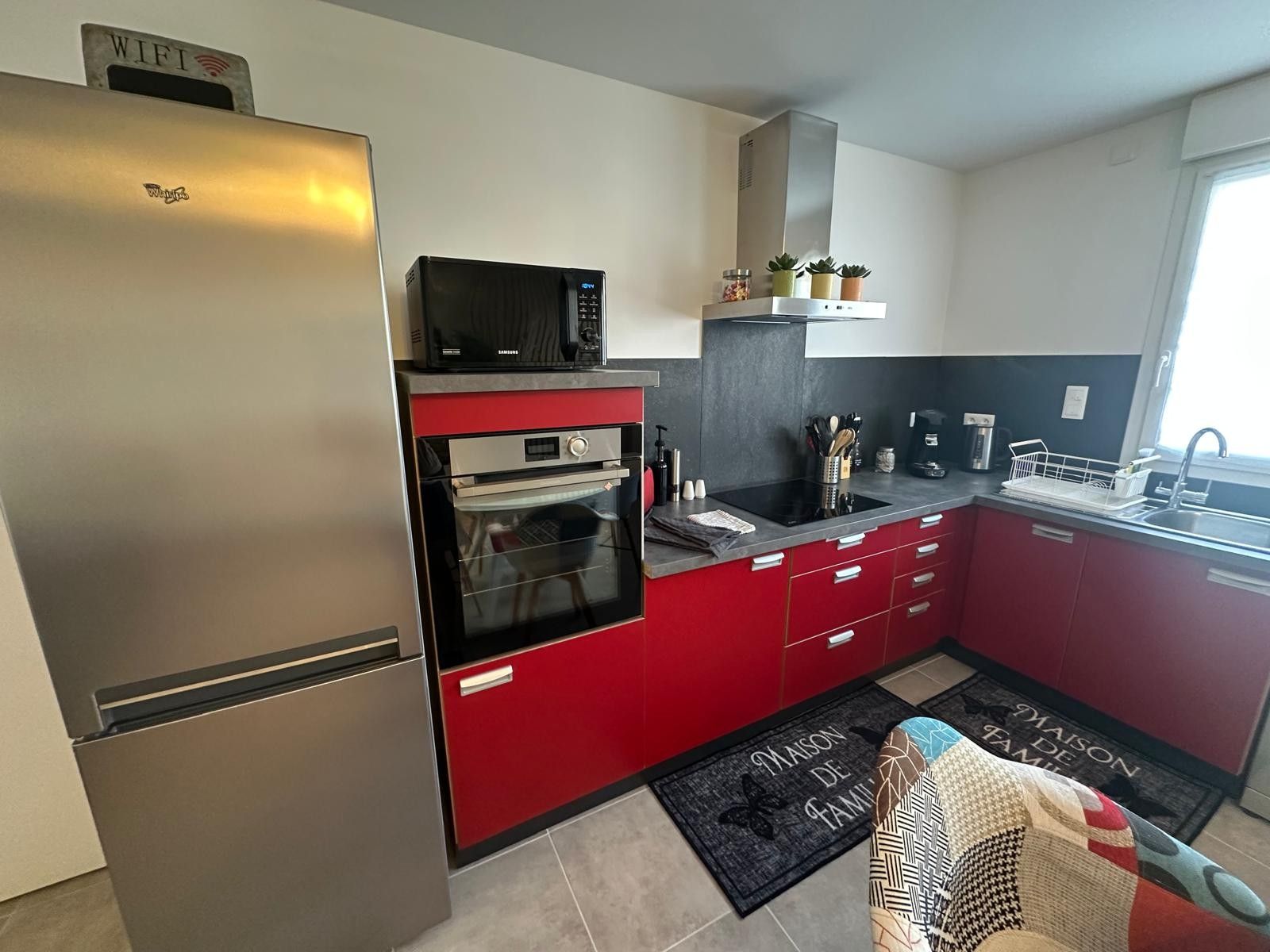 A compact kitchen with a stainless steel refrigerator, a microwave, and an oven. There is a sink with a faucet, a coffee maker, and a dish rack. The cabinets are red with silver handles.