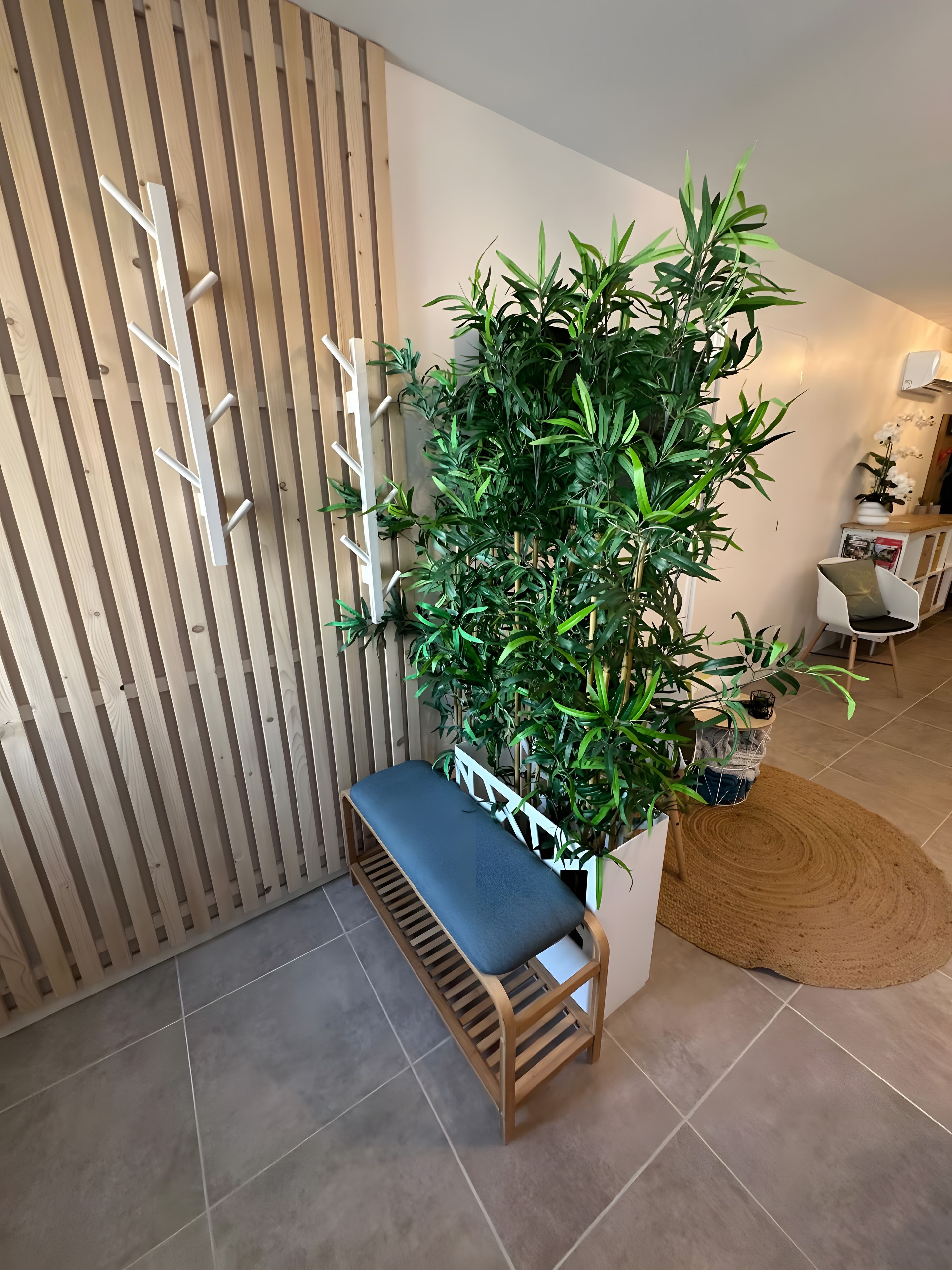 Un banc en bois avec un siège rembourré, un mur fait de lattes de bois verticales et une grande plante en pot aux feuilles vertes.