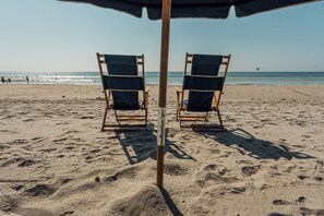 Une plage avec deux chaises de plage en bois sous un grand parasol sombre, une plage de sable et l’océan en arrière-plan.