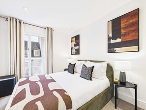 A hotel room with a bed, a nightstand, a lamp, and artwork on the walls. - Westminster 2BR Apartment with Balcony (Greater London)