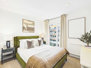 Habitación de hotel con cama, mesita de noche, lámpara, ventana con cortinas, radiador y un cuadro enmarcado en la pared.