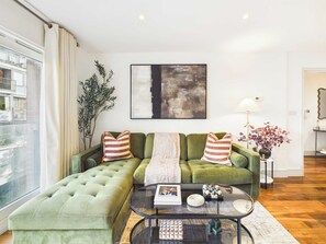 Un salon avec un canapé vert, des coussins à rayures, une table basse en verre et un tableau accroché au mur.