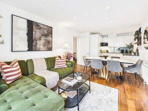 Ein modernes Wohnzimmer mit einem grünen Sofa, einem Glastisch und einem Essbereich mit einem weißen Tisch und grauen Stühlen.