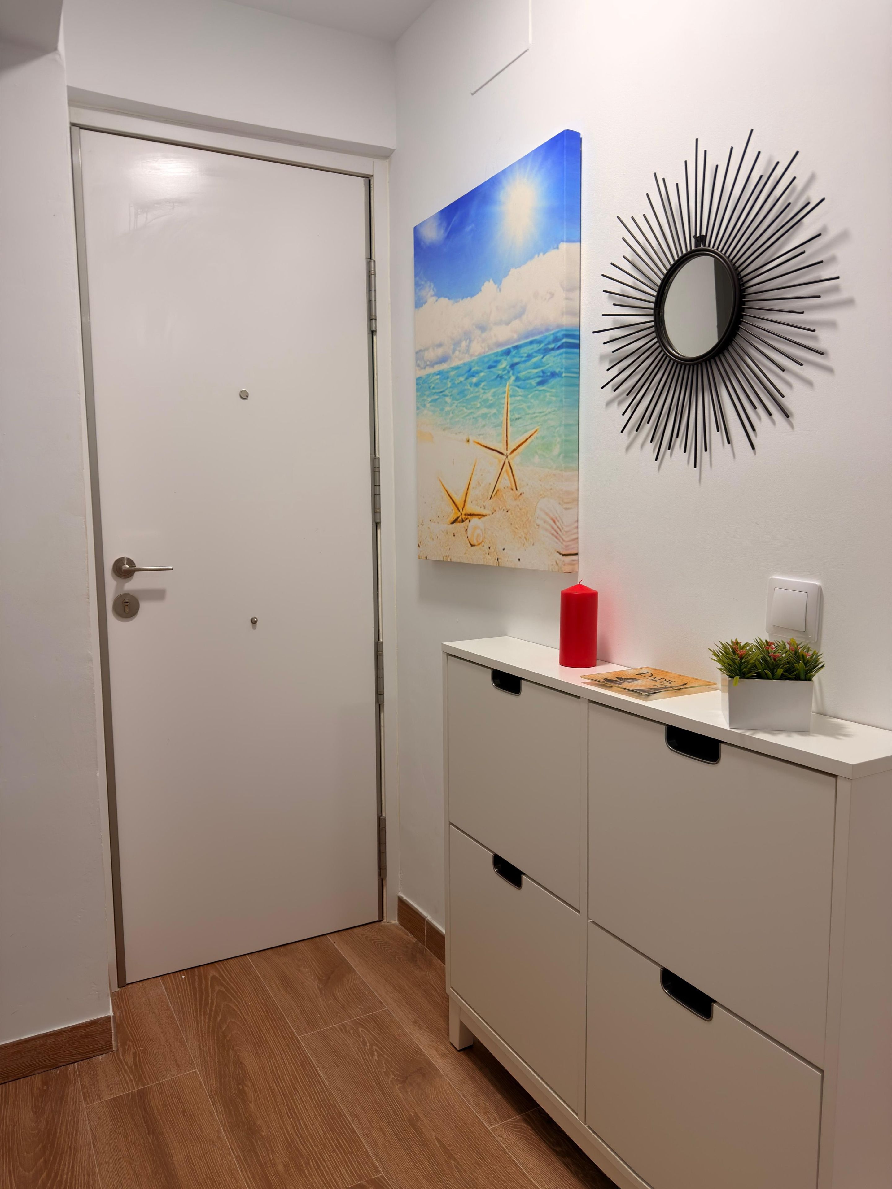 Een kamer met een witte deur, een wandspiegel, een schilderij van een strandlandschap en een wit kastje met lades.