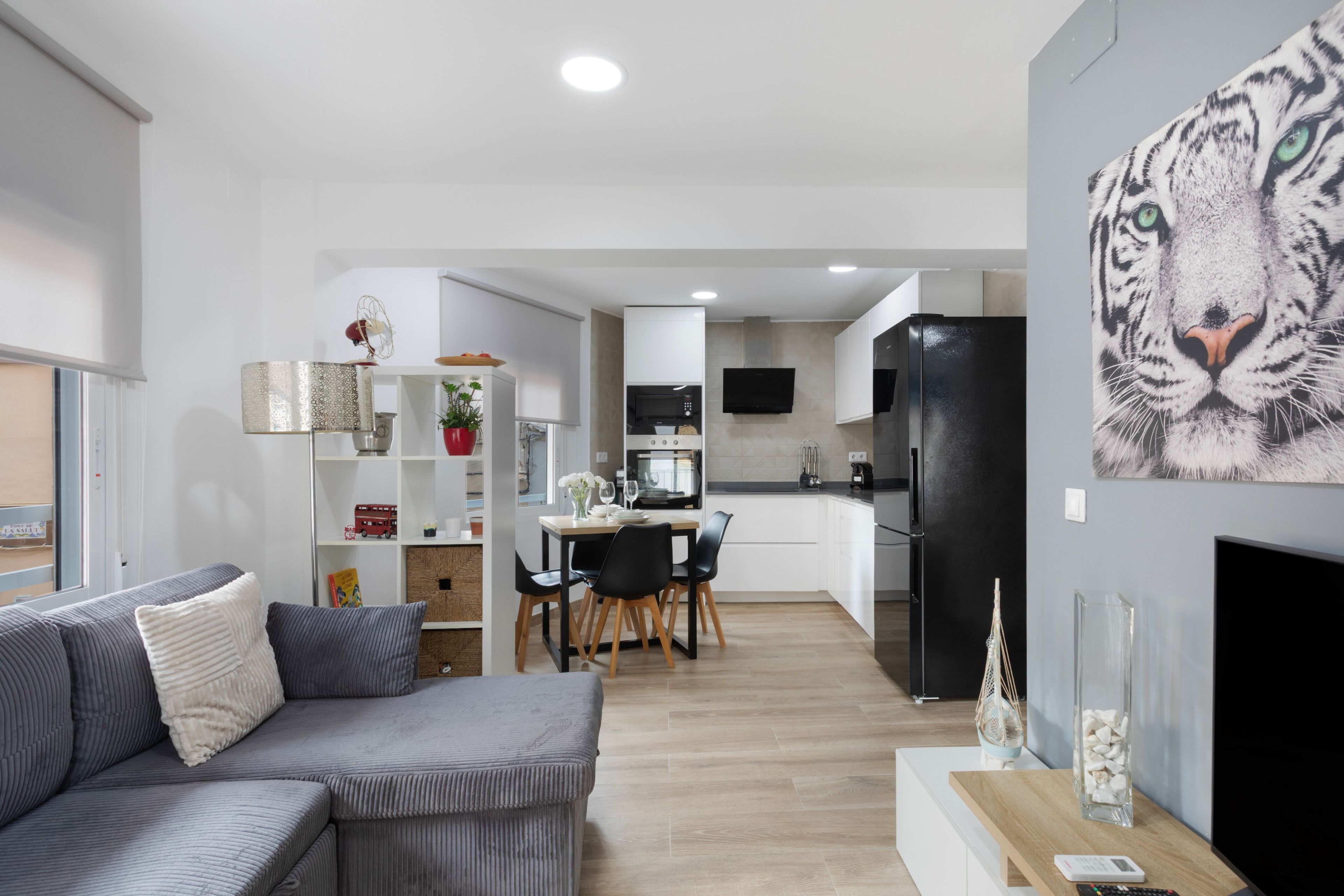 A modern living room with a grey sofa, a dining area with a table and chairs, a kitchen with a black refrigerator, and a large tiger mural on the wall.