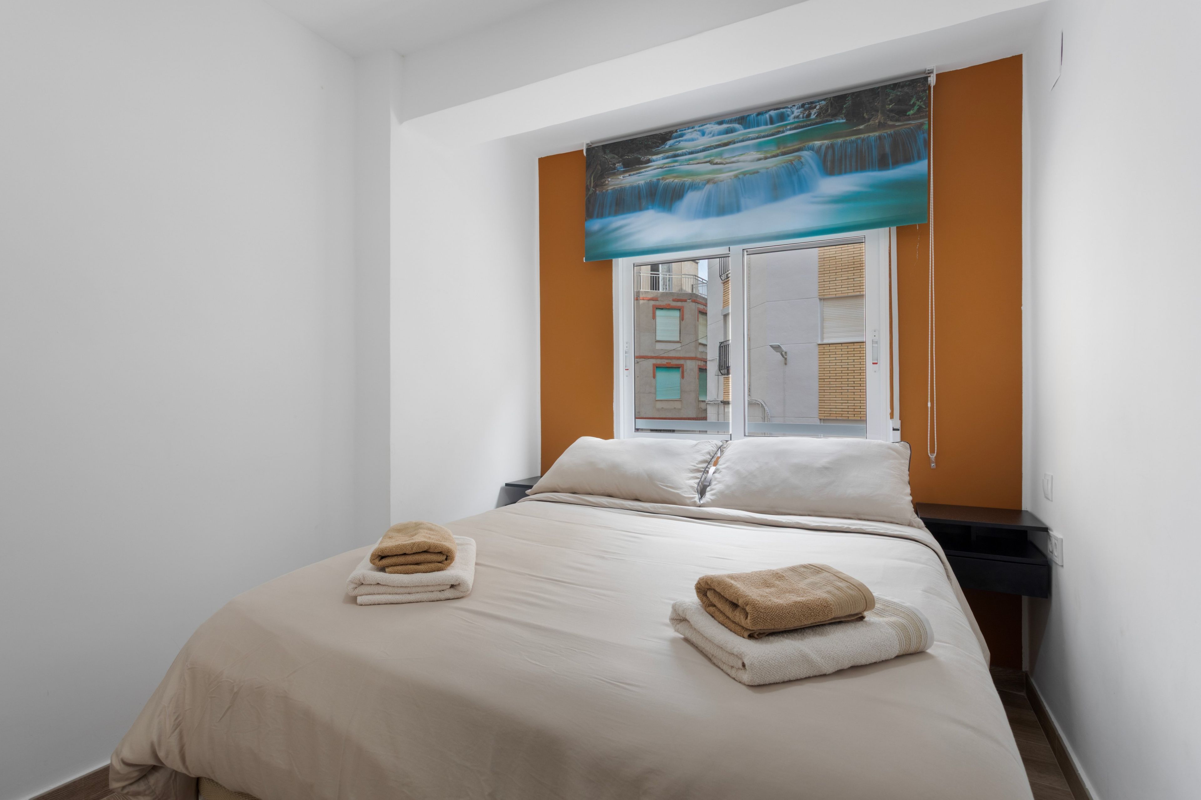 Ein Hotelzimmer mit einem Bett, zwei Handtüchern, einem Fenster mit Blick auf ein Gebäude und einem Gemälde über dem Fenster.