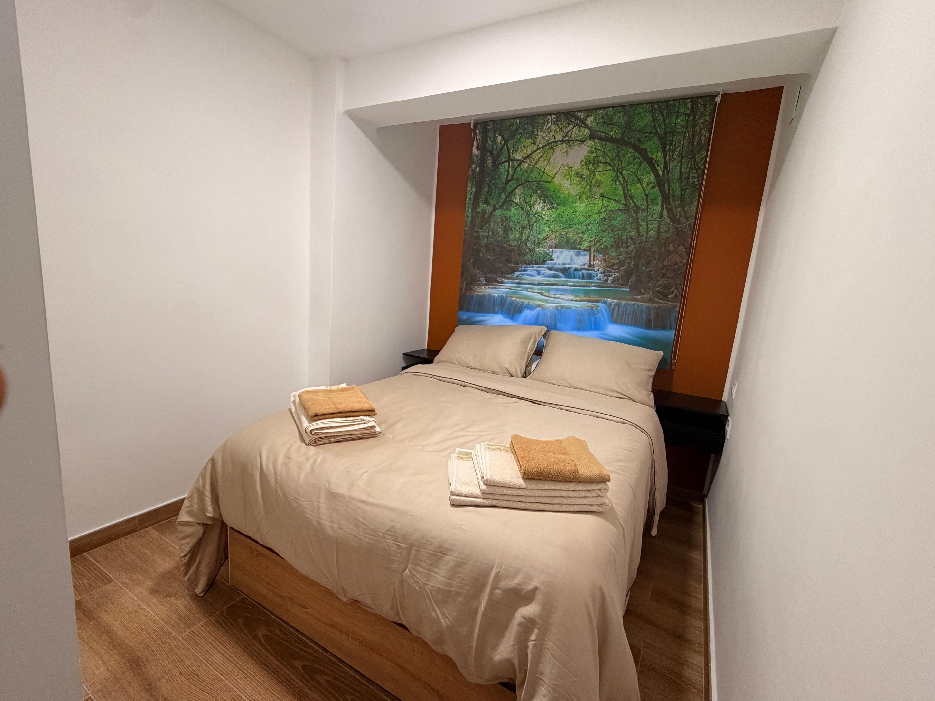 Cama bem arrumada com colcha bege e almofadas, mesa de cabeceira com abajur e um grande mural na parede retratando uma cascata em uma floresta.