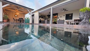 Ein Schwimmbecken mit glatter Wasseroberfläche, umgeben von einer überdachten Terrasse mit Sitzgelegenheiten und einem Deckenventilator.