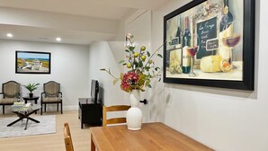 Uma área de refeições com mesa de madeira, cadeiras, um vaso com flores, um quadro de garrafas de vinho e uma fotografia emoldurada de uma praia.