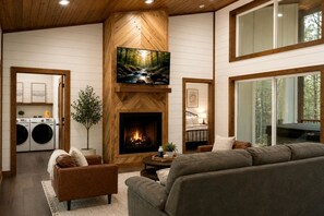 A living room with a fireplace, a television mounted above it, and a large window providing a view of the outdoors. - Luxury Cabin with Private Hot Tub & Sauna Getaway in Broken Bow, Oklahoma (Broken Bow)