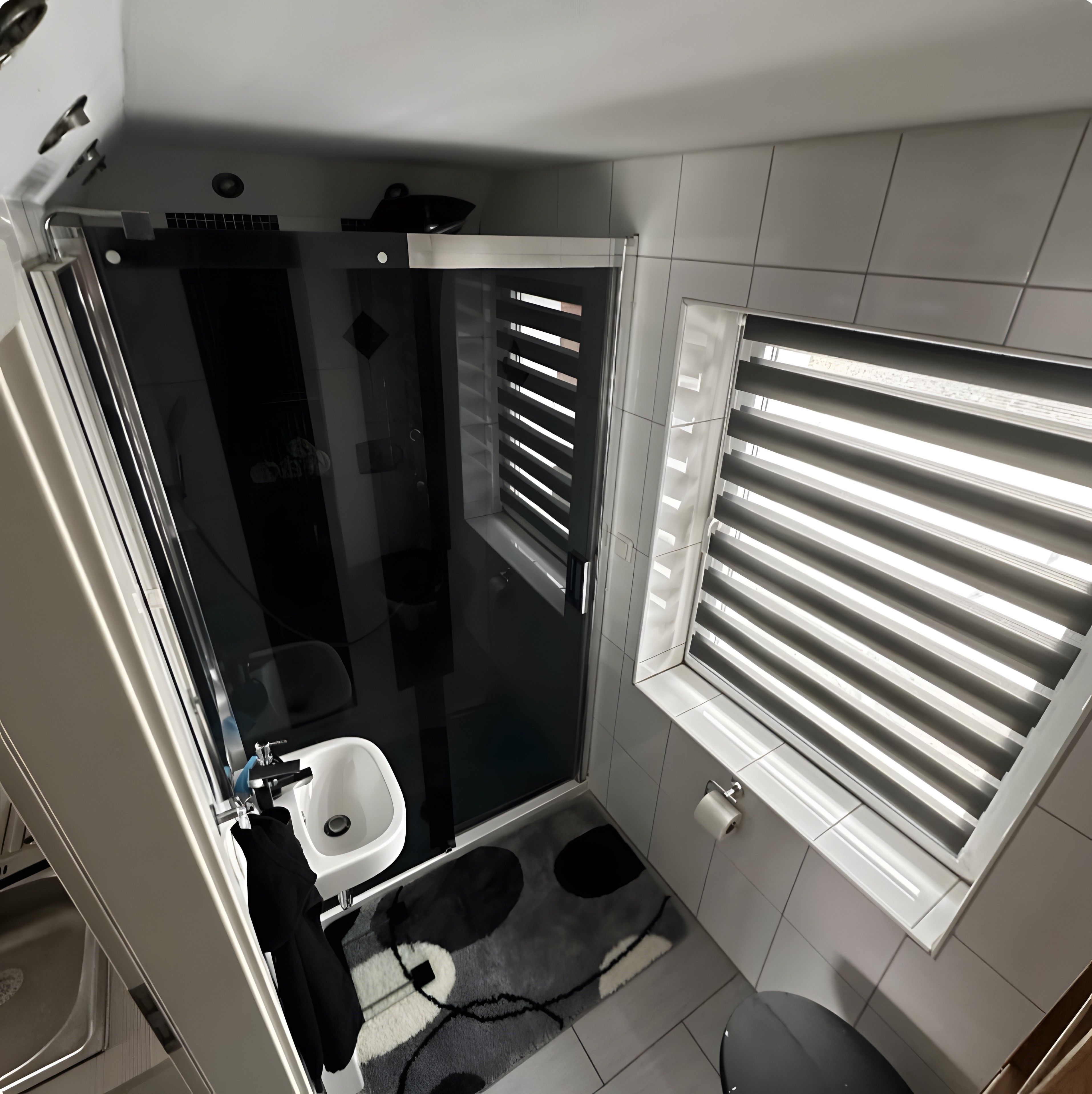 Ein kleines Badezimmer mit Glasdusche, Toilette und Fenster mit Jalousien.