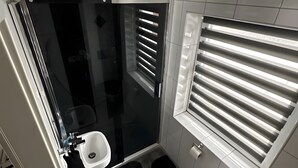 Un baño compacto con cabina de ducha de vidrio, inodoro y ventana con persianas.