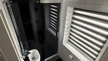 Ein kleines Badezimmer mit Glasdusche, Toilette und Fenster mit Jalousien.