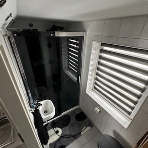 A compact bathroom with a glass shower enclosure, a toilet, and a window with blinds. - Apartment “Pool Oasis” with Pool, Terrace, and Wi-Fi (Colbitz)