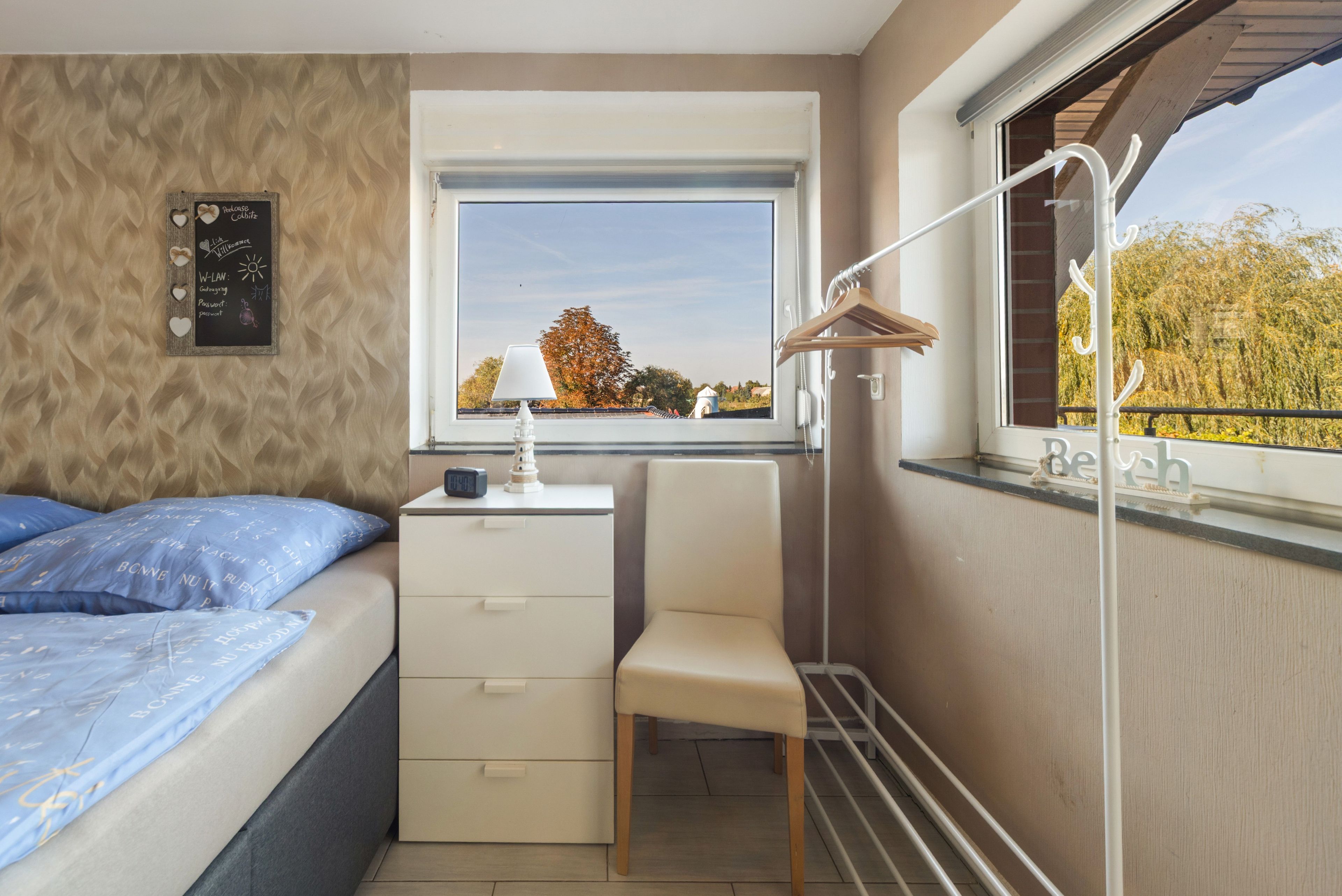 Ein Schlafzimmer mit einem Bett, einem Schreibtisch, einem Stuhl, einem Fenster mit Blick auf Bäume und einem an der Wand befestigten Regal.