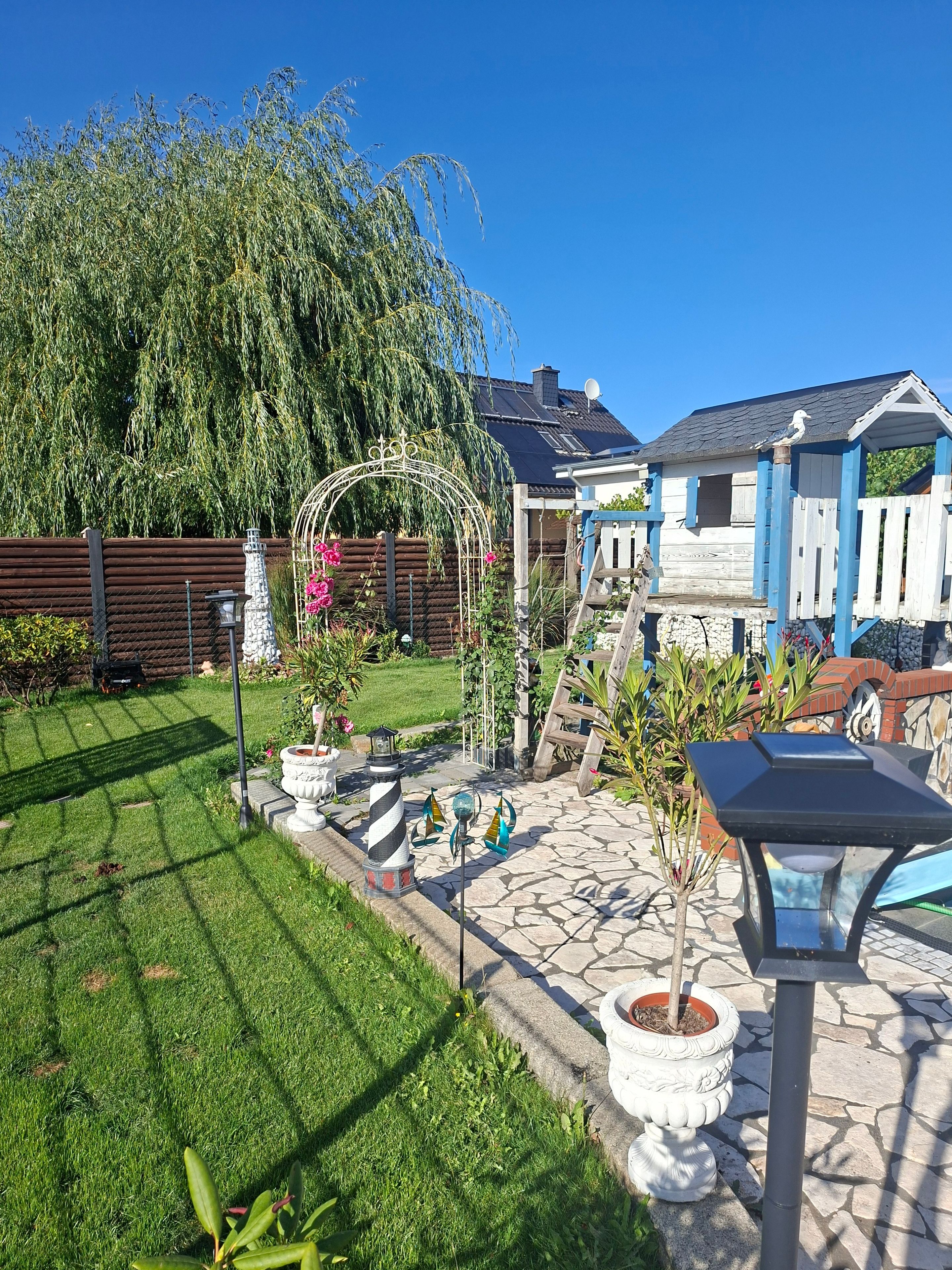 Ein gepflegter Garten mit Laube, Steinweg und verschiedenen Pflanzen.
