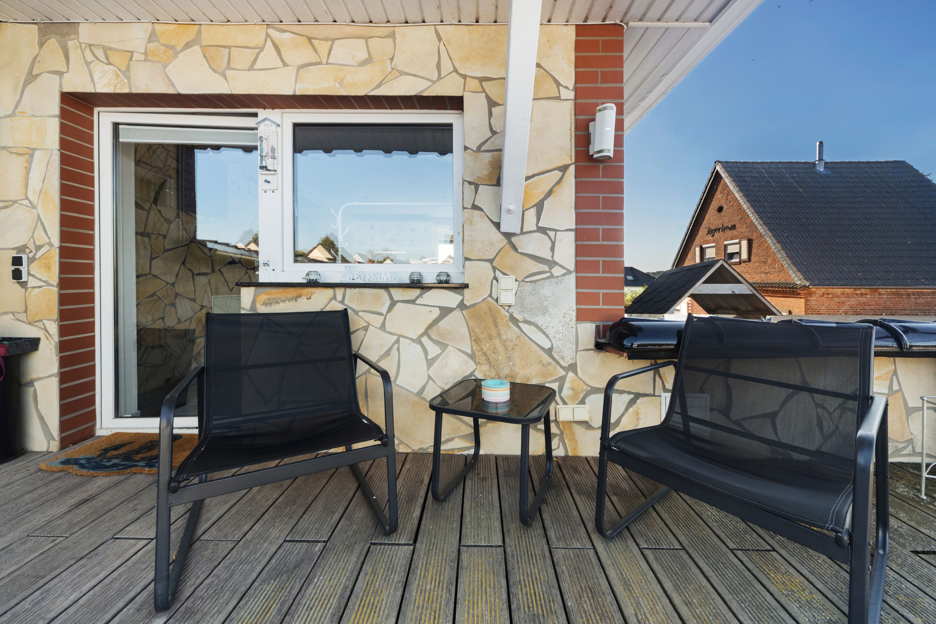 An outdoor patio with two black mesh chairs, a small table, and a stone wall with a welcome sign.