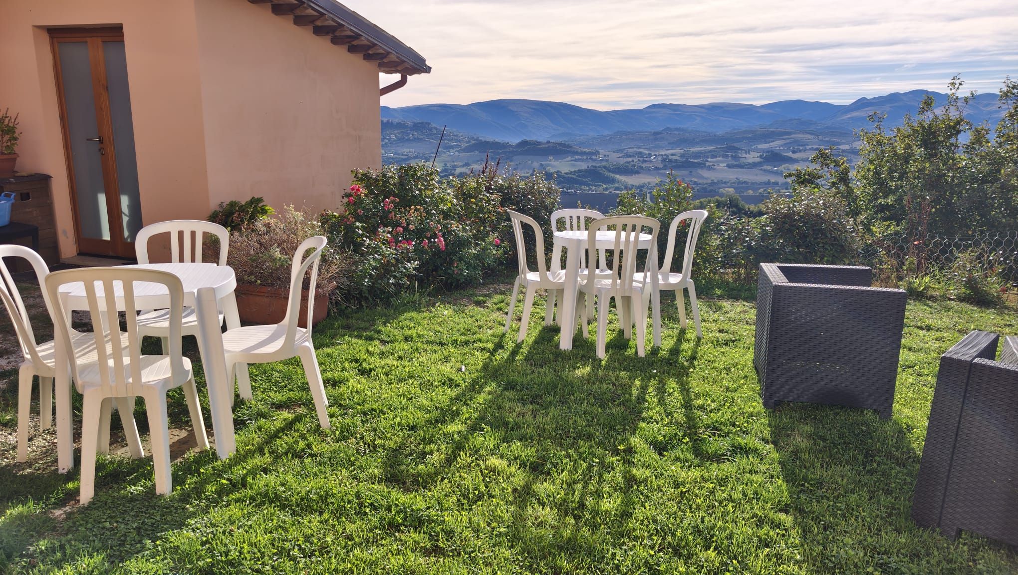 A patio with white plastic chairs and a table, surrounded by lush green grass and flowering plants, overlooking a mountainous landscape.