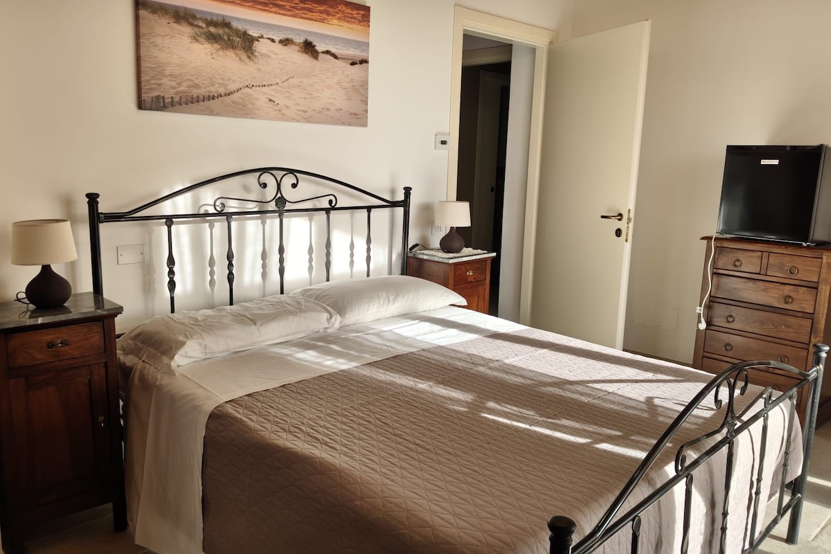 A bedroom with a bed, bedside tables, a dresser, a television on top of the dresser, and a painting on the wall.