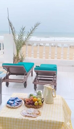 A beachside lounge area with two sun loungers, a table with fruit and pastries, and a view of the ocean.