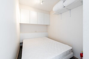 A small, white, single-bed room with a simple layout, including a bed, a white wardrobe, and a ceiling-mounted shelf. - Studio Apartment With Sea Views in Gadeynehelling (Blankenberge)