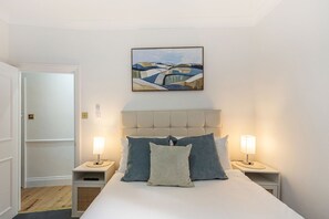 Quarto de hotel com cama, duas luminárias de cabeceira, mesa de apoio, pintura na parede e uma porta.