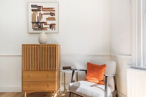 Ein Zimmer mit einem Holzkabinett, einem Sessel mit orangefarbenem Kissen und einem gerahmten abstrakten Kunstwerk an der Wand.