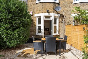 A corner bay window with a view of the garden, a wooden fence, and a patio table with chairs. - GuestReady - Charming Garden Retreat in London (London)