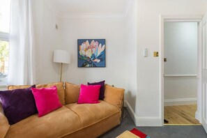Ein Wohnzimmer mit einem beigefarbenen Sofa, bunten Kissen, einer Stehlampe und einem Bild an der Wand.