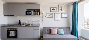 Eine kompakte Küche mit Einbauofen, Kaffeemaschine und Mikrowelle. Es gibt ein graues Sofa mit Kissen und eine Stehlampe.