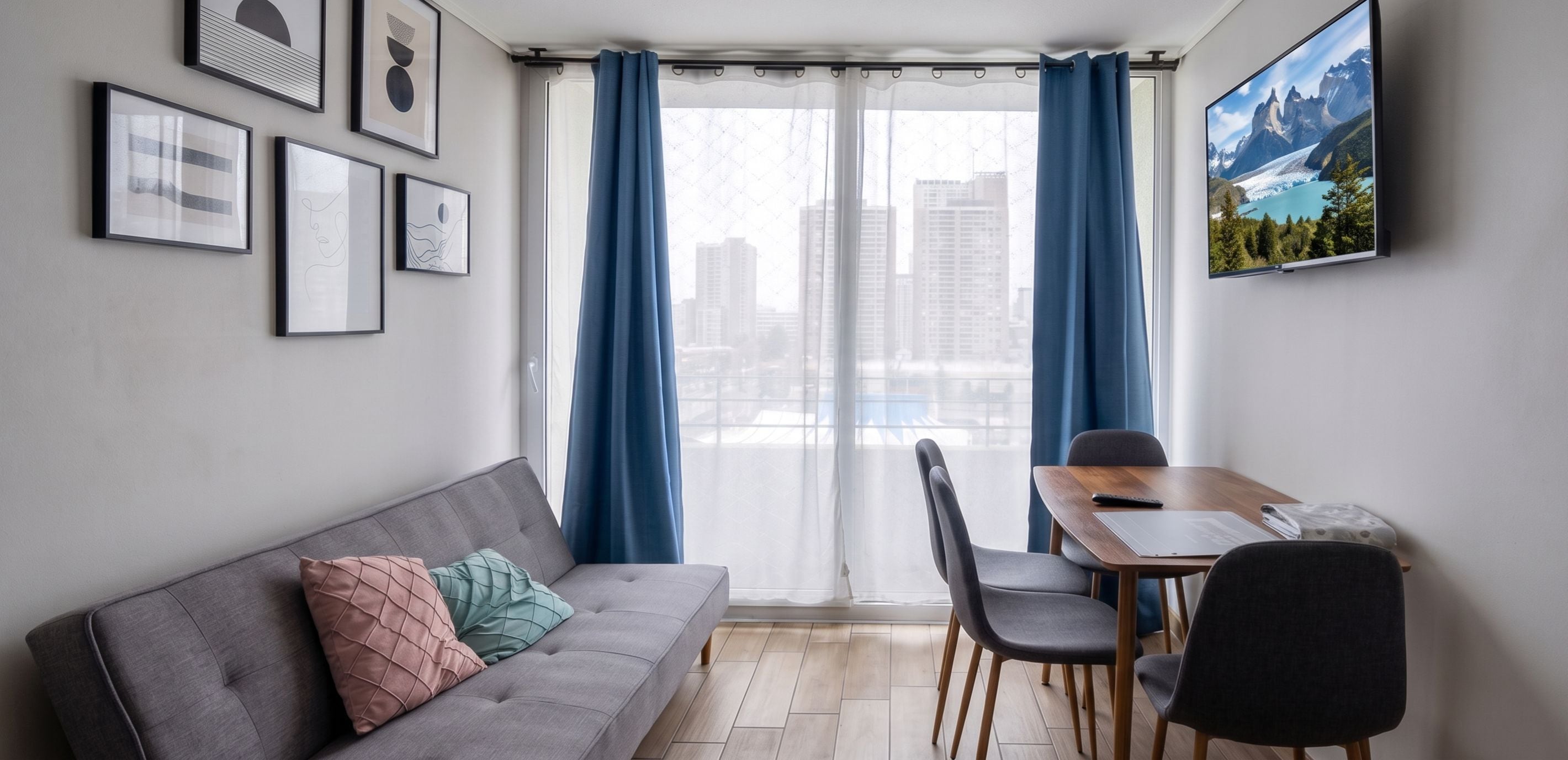 Una sala de estar moderna con un sofá gris, una mesa de comedor con sillas, vistas urbanas a través de ventanas amplias y obras de arte enmarcadas en la pared.