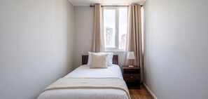 Ein schmales Hotelzimmer mit Bett, Nachttisch, Lampe und einem Fenster mit Vorhängen.