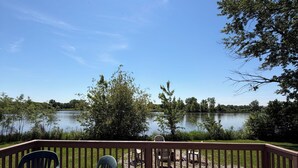 A wooden deck with a table and chairs, overlooking a calm lake and surrounded by trees. - Shoreline Haven (Erie)