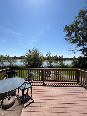 Une terrasse en bois avec une table et des chaises, donnant sur un lac paisible et entourée d’arbres.