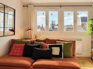 Ein Wohnzimmer mit einer Couch, Kissen, einer Lampe und Blick auf Gebäude durch Fenster.