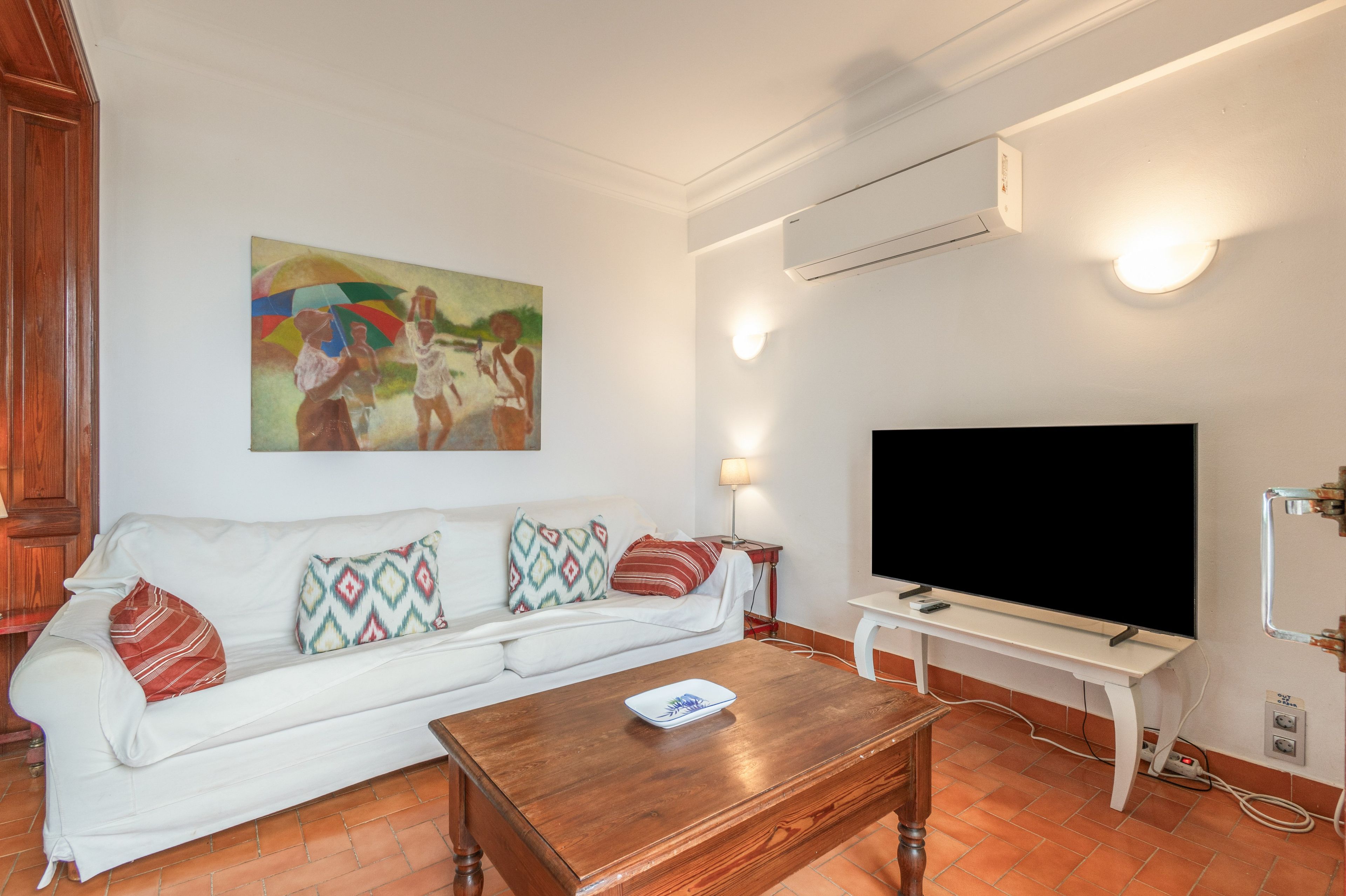 Ein Wohnzimmer mit einem weißen Sofa, gemusterten Kissen, einem Holztisch, einem Flachbildfernseher und einer Klimaanlage.