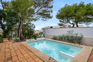 A swimming pool with a tiled deck, surrounded by greenery and a white wall.